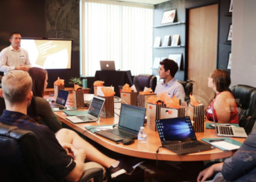 A presenter speaks to attendees gathered around a conference table during a meeting.
